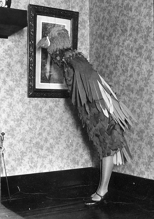 Vintage Halloween costume of a person dressed as a large bird looking at a framed picture on a wallpapered wall.