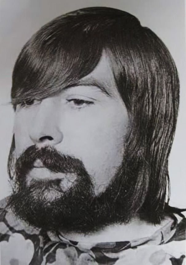 Man with 1970s hairstyle, featuring long hair and a full beard, in a black and white photo.