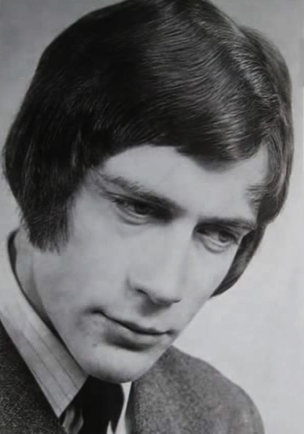 Man with 1970s romantic hairstyle, featuring sleek, side-parted hair and sideburns, dressed in a suit.