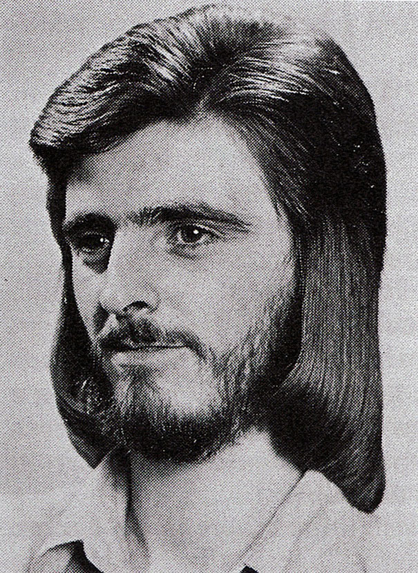 Man with long 1970s hairstyle and beard, showcasing retro men's fashion.