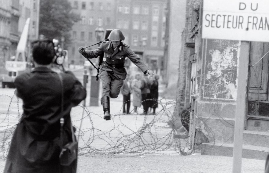 Soldier jumping over a barbed wire barrier during the Berlin Wall construction, captured in a famous historical photo.