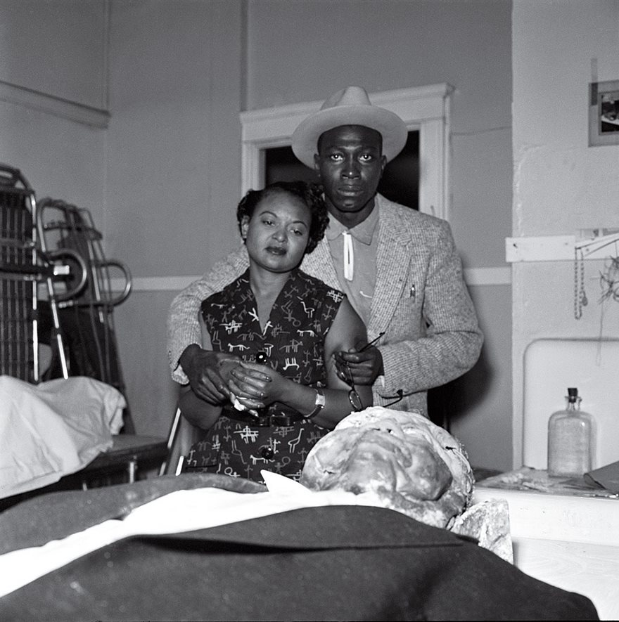 A historical photo of a couple in a somber setting, depicting a poignant moment with an embalmed body in the foreground.