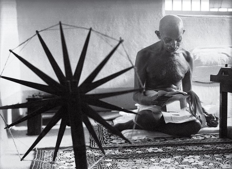 A historical figure, seated next to a spinning wheel, reading in a simple room.