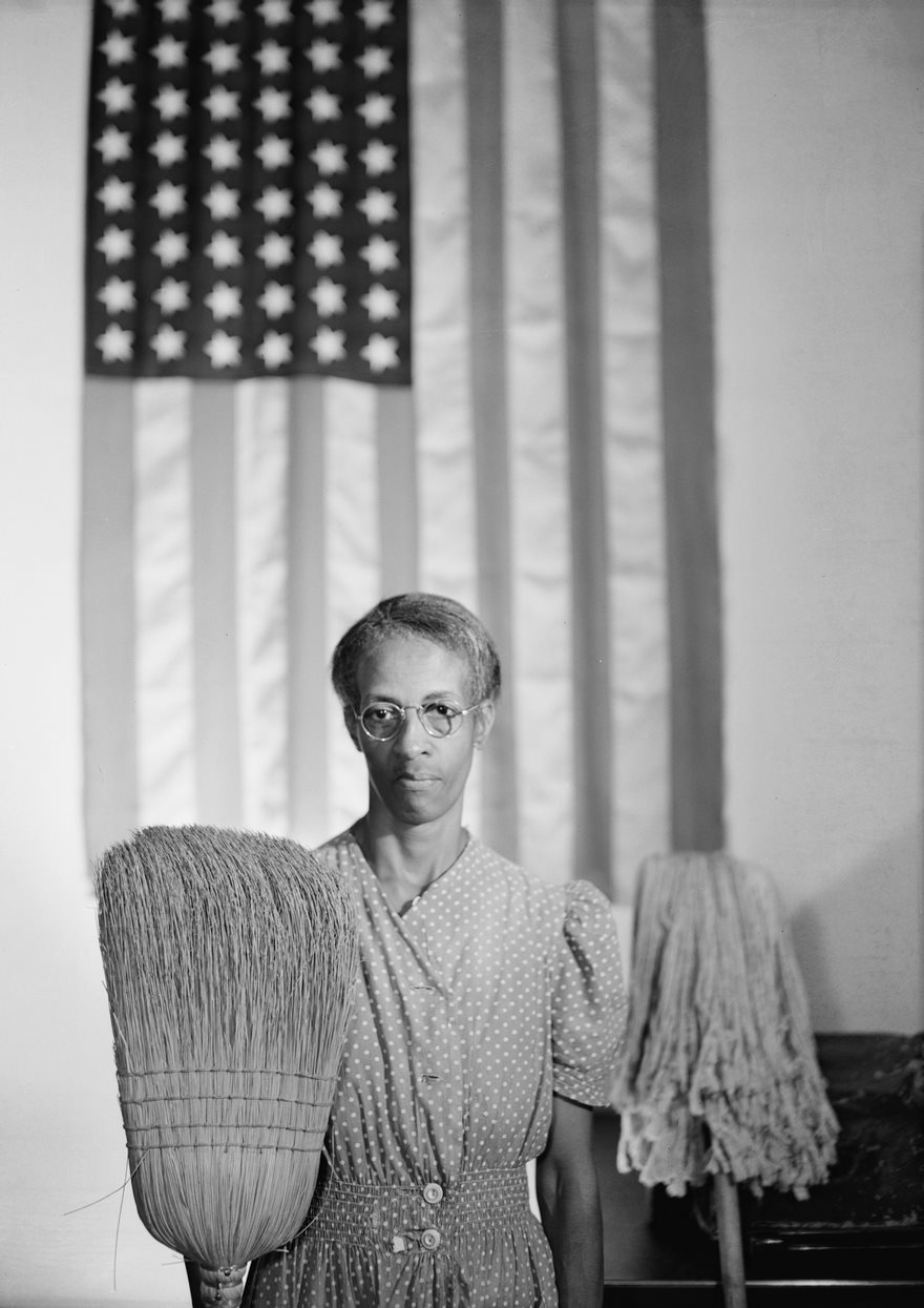 A woman in glasses holding a broom stands in front of an American flag, representing a famous historical photo.