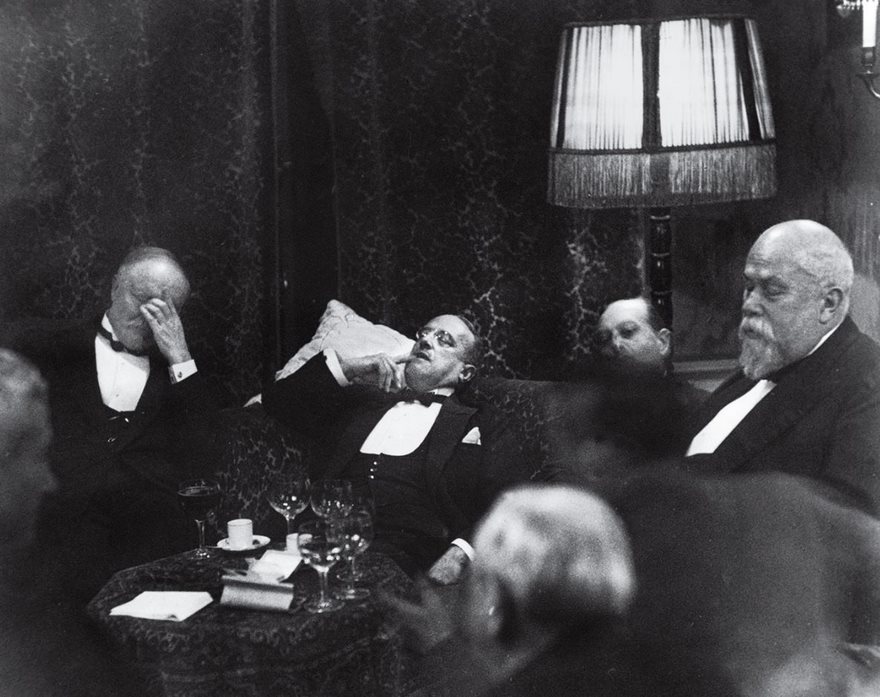 A historical black and white photo depicts four men in suits relaxing in a dimly lit room, conveying a classic era ambiance.