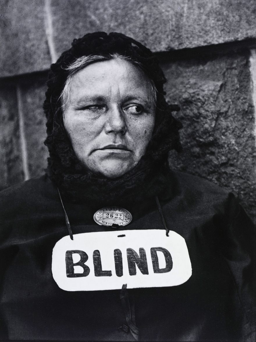 Woman in a headscarf with "BLIND" sign, from a famous historical photo, against a textured wall.