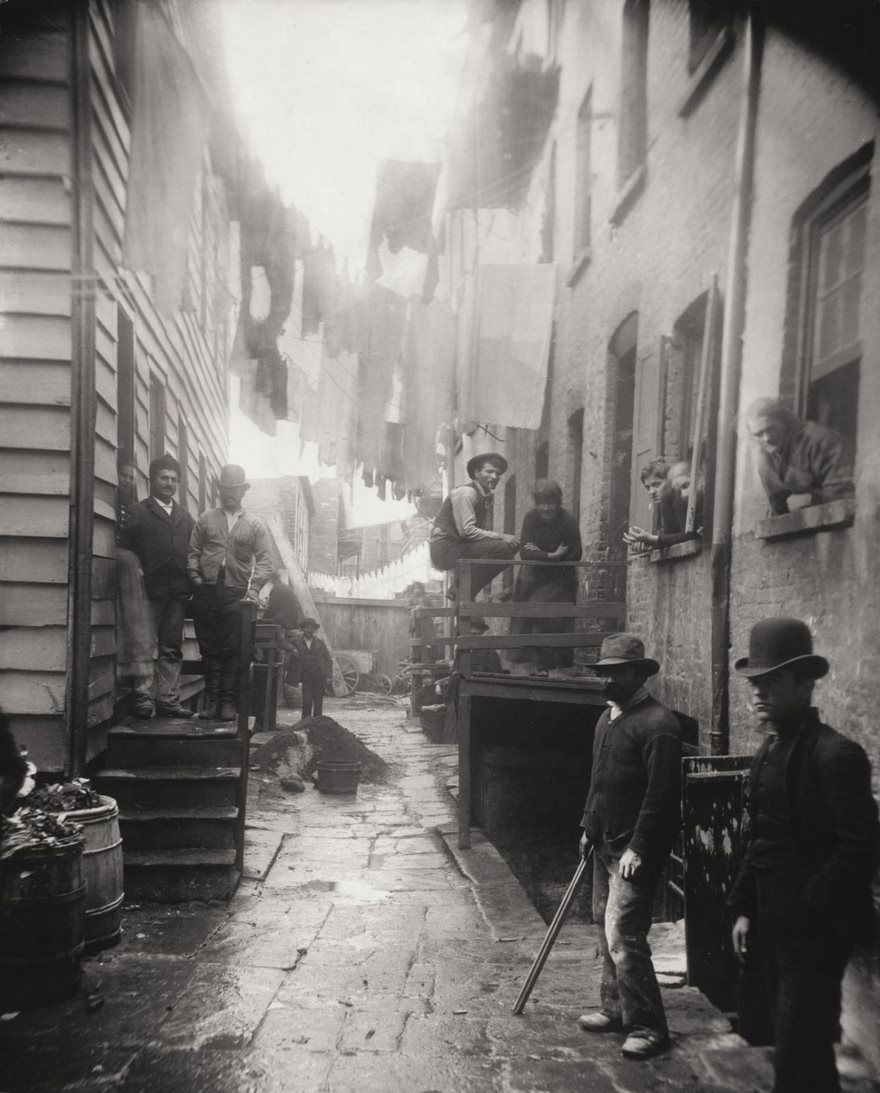 Historic photo of people in a narrow alley, wearing hats and traditional clothing, with laundry hanging above.