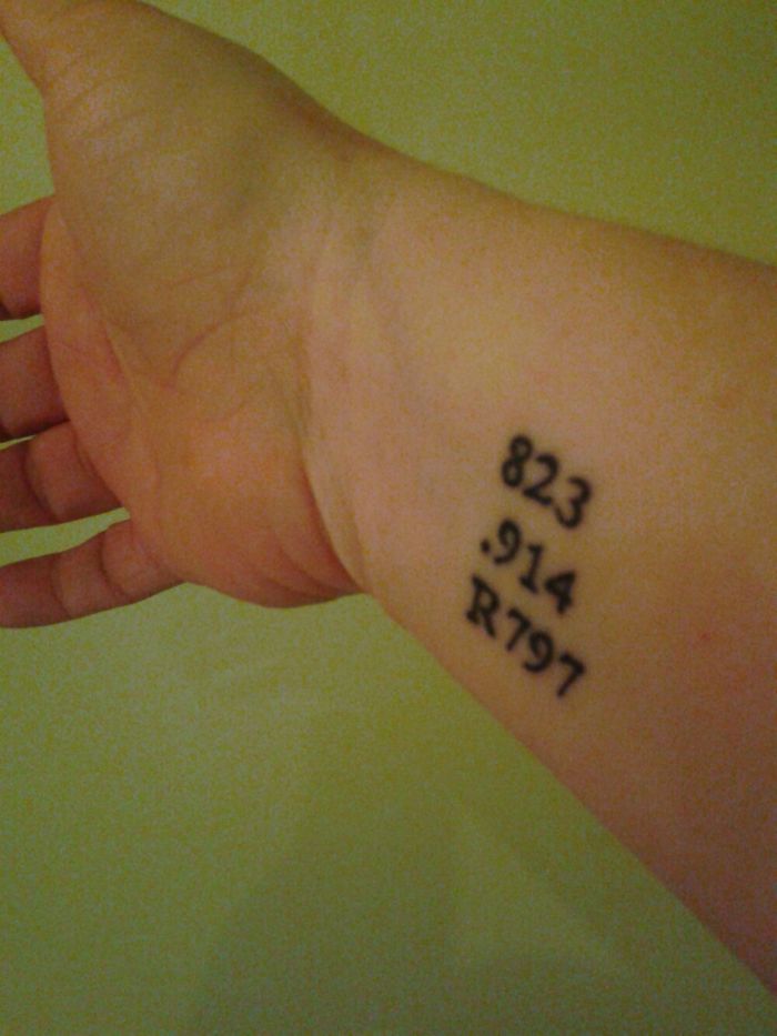 Subtle Harry Potter tattoo on wrist with numbers 823.914 R797 in black ink.