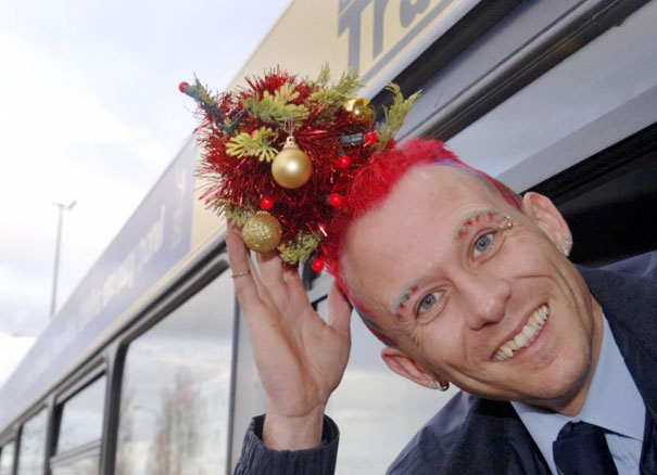 Man with red hair styled as a creative Christmas tree, featuring ornaments and lights, smiling from a bus window.