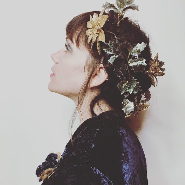 A woman with a creative Christmas hairstyle featuring gold flowers and green leaves, wearing a dark velvet outfit.
