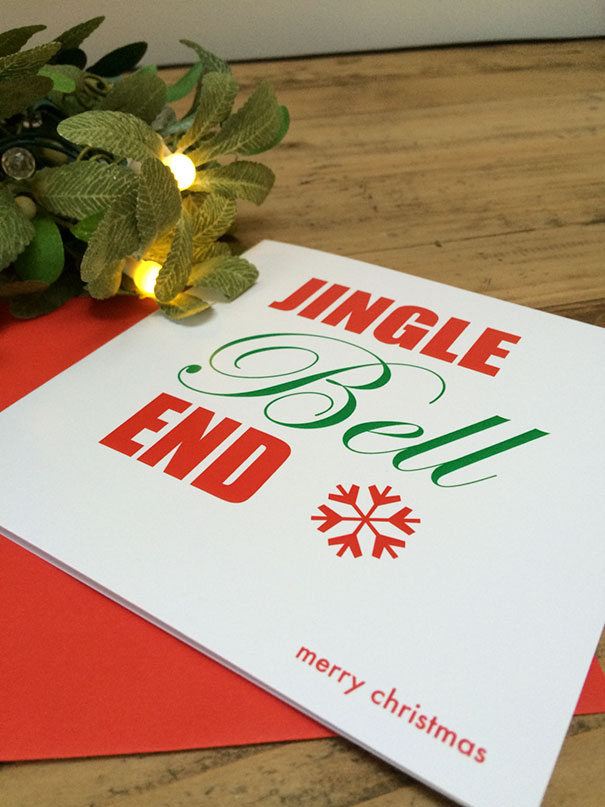 Rude Christmas card with "Jingle Bell End" text, showcasing twisted humor with festive decor on a wooden table.
