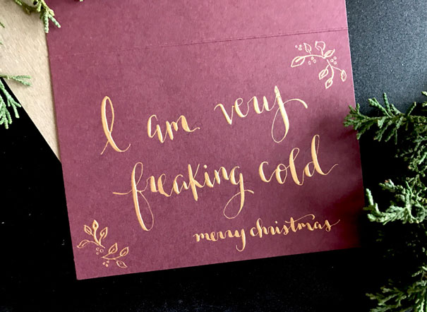 Rude Christmas card with gold lettering on a burgundy background saying "I am very freaking cold, merry christmas."