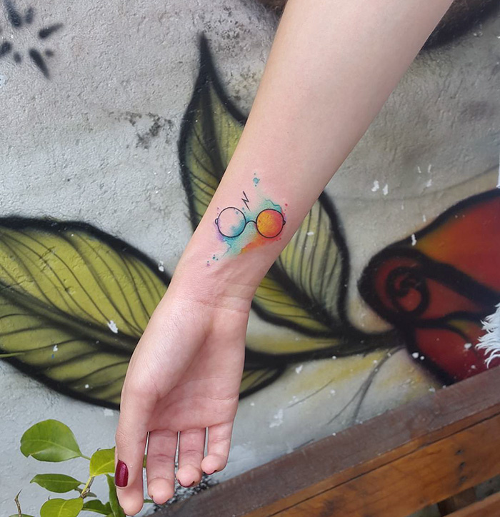 Colorful subtle Harry Potter tattoo featuring glasses and a lightning bolt on a forearm, set against a floral mural background.