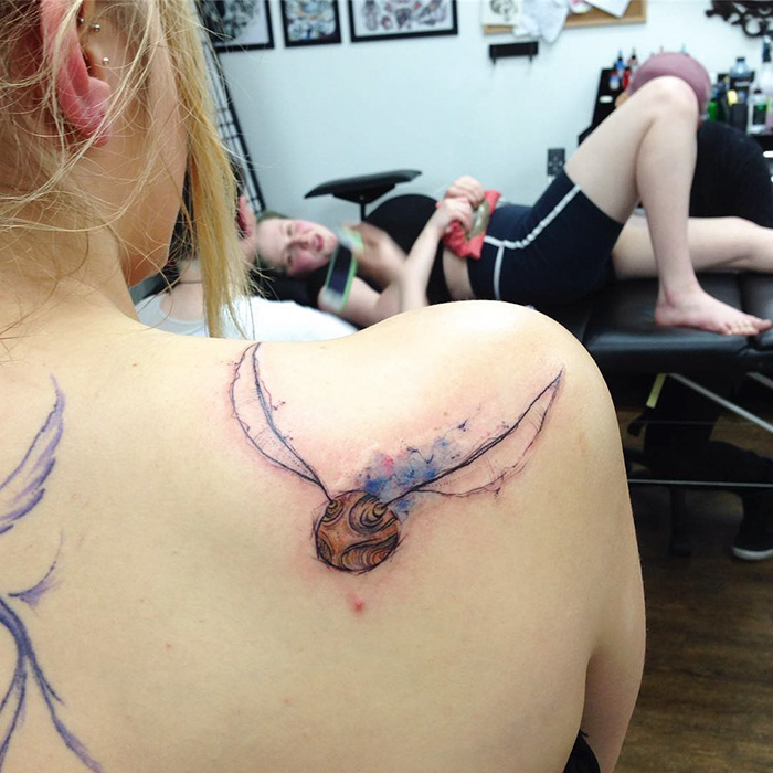 Golden Snitch tattoo on woman's shoulder at a tattoo parlor; subtle Harry Potter design for true fans.