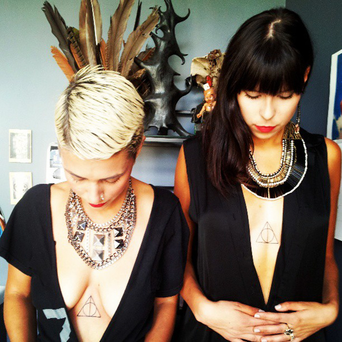 Two people displaying subtle Harry Potter tattoos on their chests, with Deathly Hallows symbols, wearing stylish black outfits.