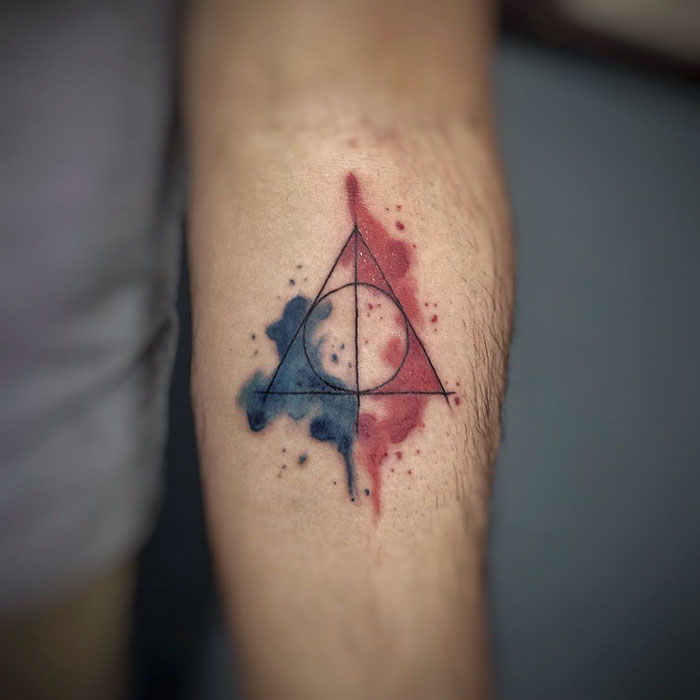 Subtle Harry Potter tattoo of the Deathly Hallows symbol with red and blue watercolor background on an arm.