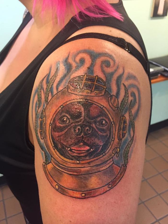 Dog tattoo of a pug in a deep-sea diving helmet on a person's arm with pink hair in the background.