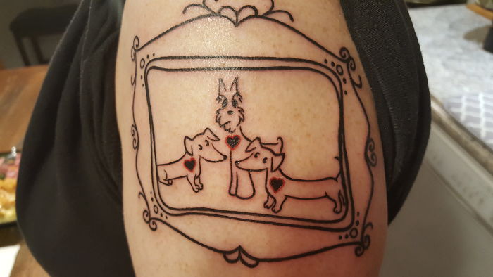 Dog tattoo featuring three outlined dogs with small hearts, framed with decorative lines on a person's upper arm.