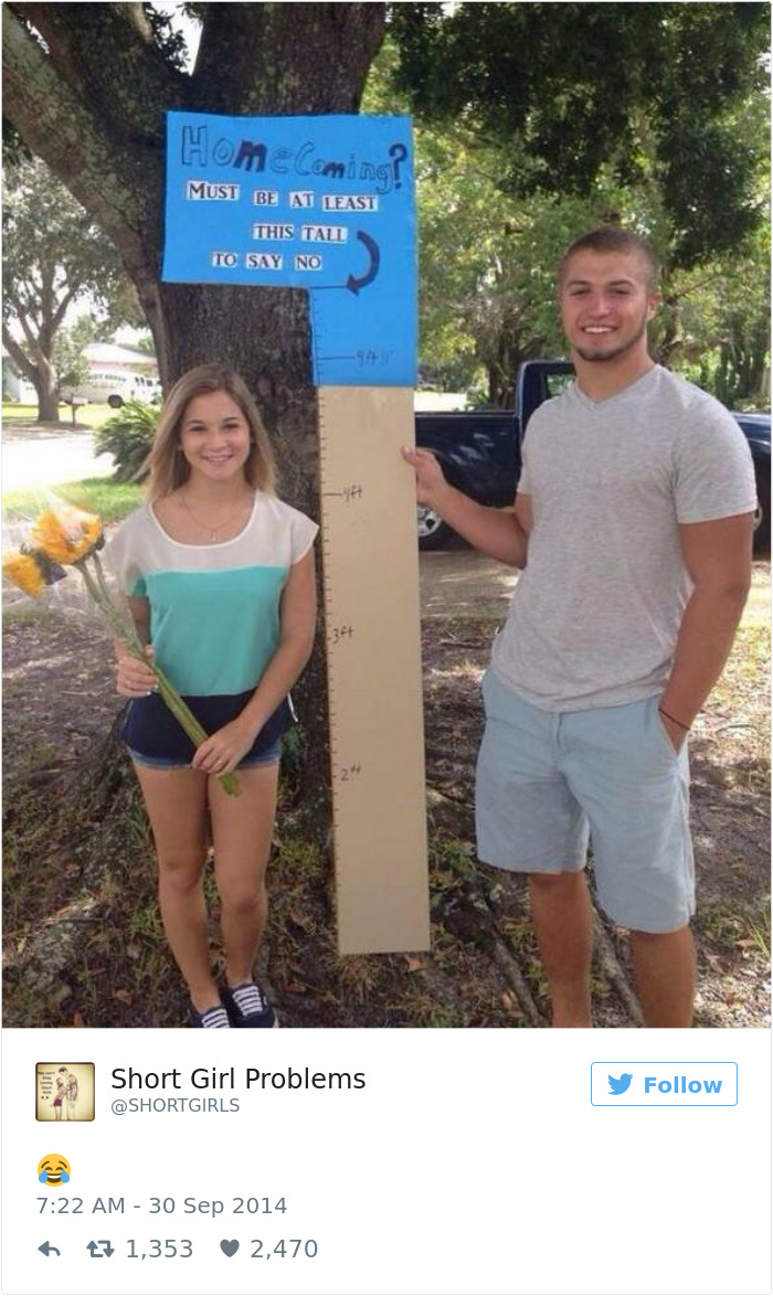 Short girl with flowers next to a tall guy holding a height requirement sign for homecoming under a tree.