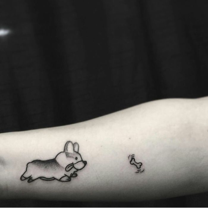 A small dog tattoo featuring a playful cartoon corgi chasing a bone on a person's forearm.