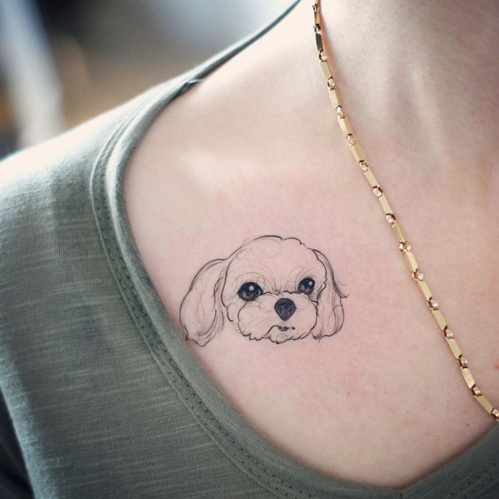 Dog tattoo on person's shoulder with fine line art style, featuring a cute puppy face above a green shirt neckline.