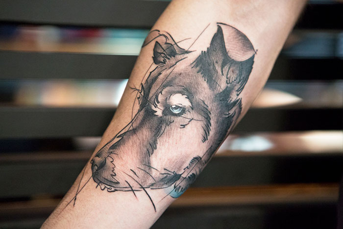 Dog tattoo design sketched on forearm, featuring a detailed canine face in grayscale ink.