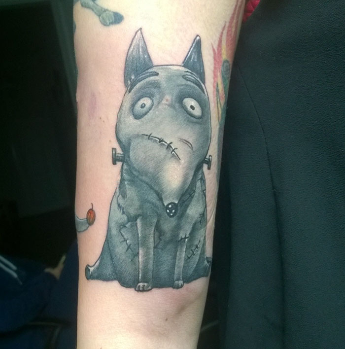 A unique dog tattoo design featuring a stitched, cartoonish canine with a whimsical expression on the arm.