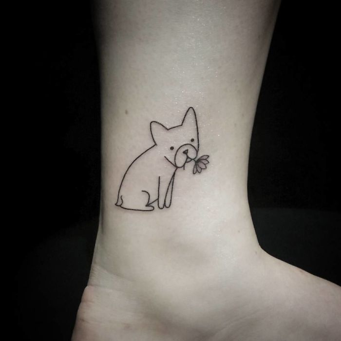 Minimalistic dog tattoo on ankle with a flower in its mouth.