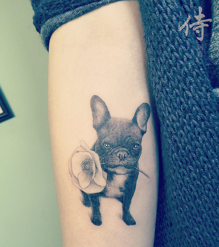Dog tattoo of a small French bulldog holding a flower, inked on an arm.