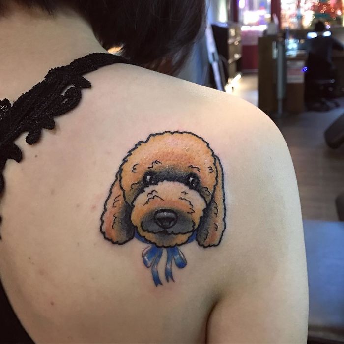 Dog tattoo on shoulder featuring a fluffy dog with a blue ribbon, highlighting unique dog tattoo ideas.