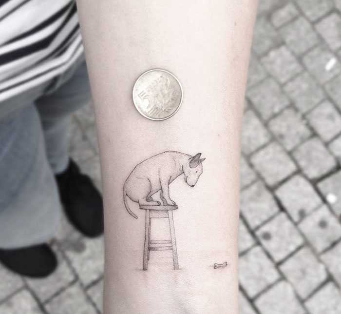 Minimalist dog tattoo of a canine on a stool, next to a coin, on a forearm showing creative design ideas.