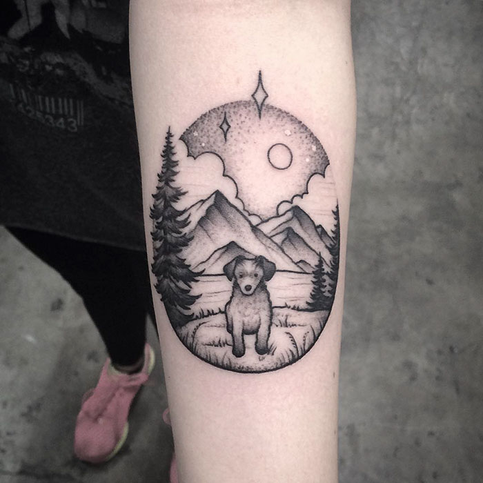 Dog tattoo with a scenic mountain and forest design on forearm.
