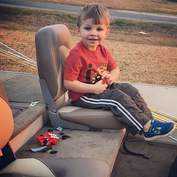 Child with home haircut sitting on a boat seat, wearing a red shirt and gray pants, with hair clippings on the floor.