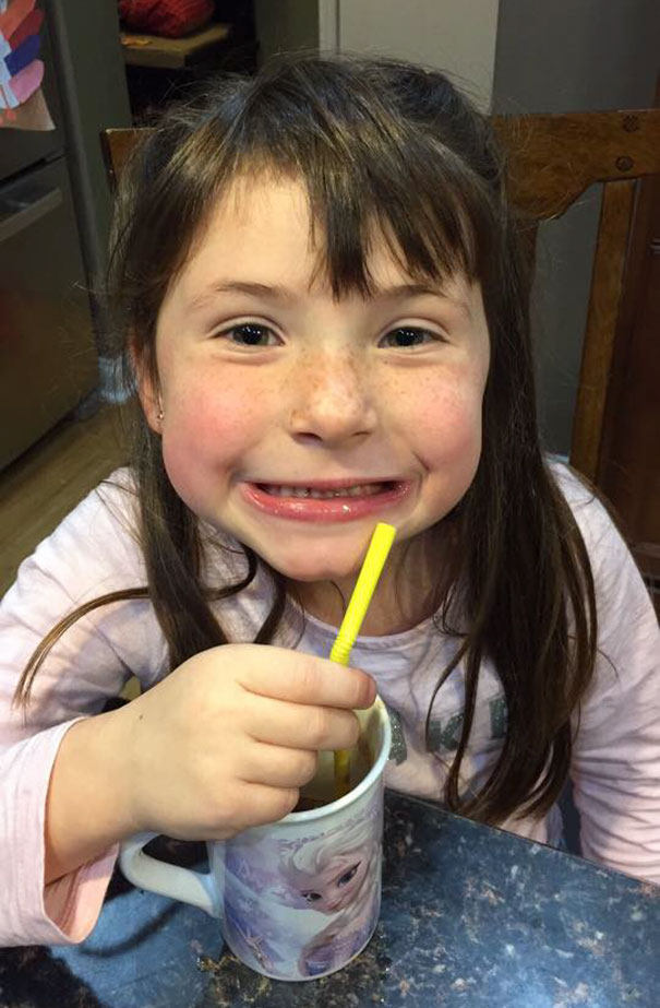Young girl with uneven bangs, sipping from a mug with a yellow straw, smiling after cutting her own hair.