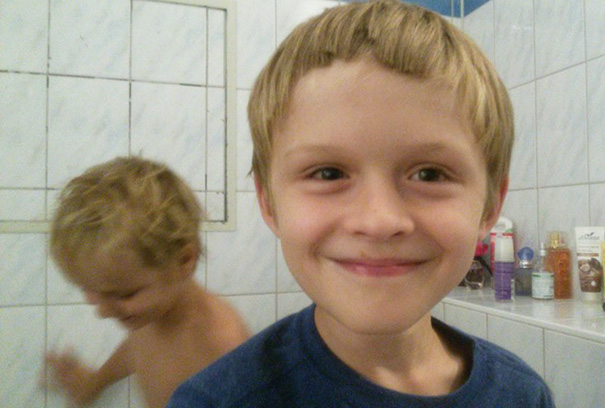Child with a self-cut haircut smiling in a bathroom, another child in the background.