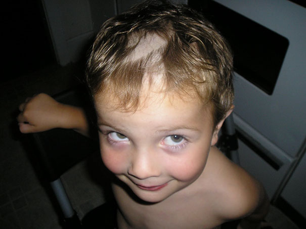 Child with uneven self-cut hair, revealing a bald spot, smiling cheekily up at the camera indoors.