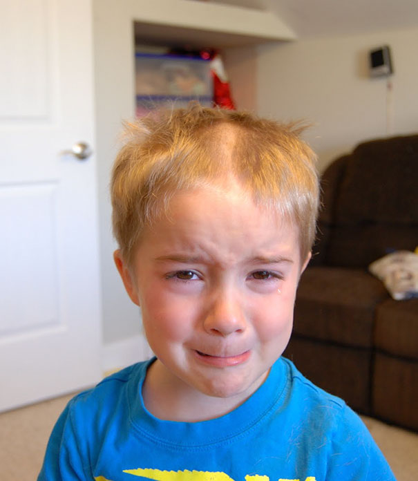 A child with a self-inflicted haircut looks upset, standing in a room.