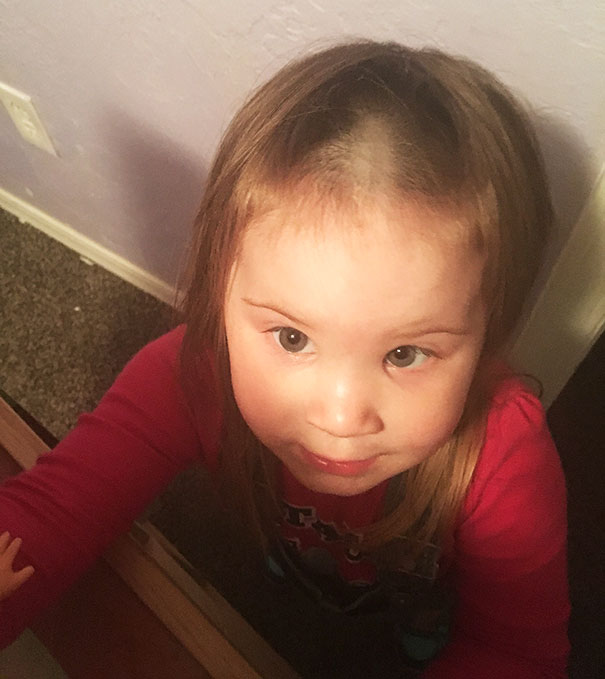 Child with a self-cut hairstyle looking up, wearing a red shirt, inside a room with a light purple wall.