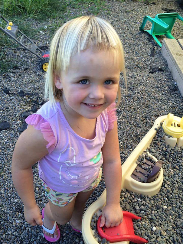 Child with a self-cut hairstyle, wearing a purple top, smiling in a gravel play area.
