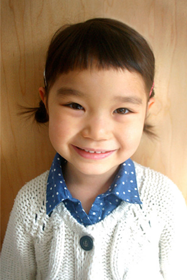Smiling child with a self-cut uneven fringe, wearing a blue polka dot shirt and white knitted sweater.