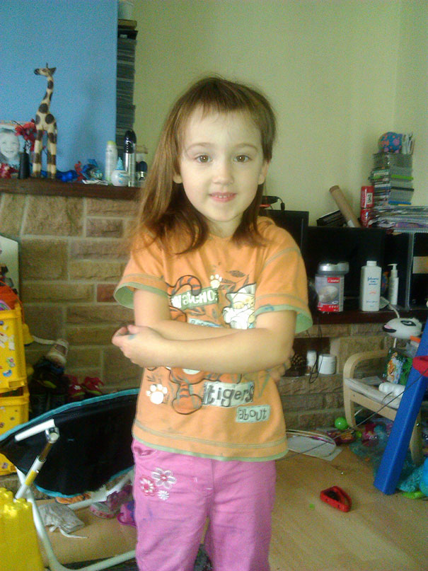 Child with a self-cut hairstyle, wearing an orange shirt, standing in a playroom setting.