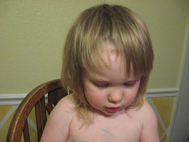 Child with self-cut uneven hair, sitting on a chair, and looking down.