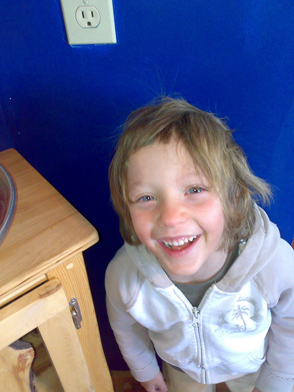 Child smiling with uneven self-cut hair, wearing a hoodie, standing near a wooden table against a blue wall.