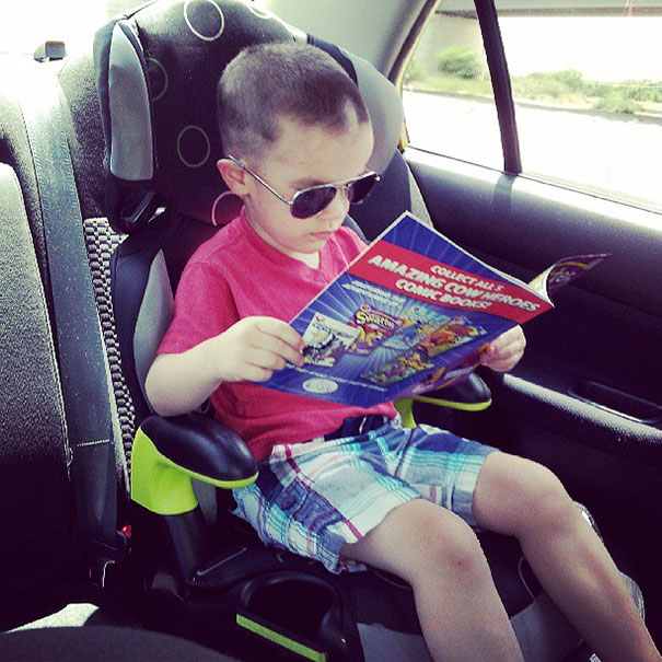Young child sits in a car seat wearing sunglasses, holding a comic book, with self-cut hair visible.