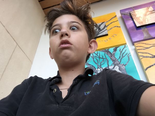 Child with a surprised expression after cutting their own hair, standing indoors with colorful artwork on the walls.