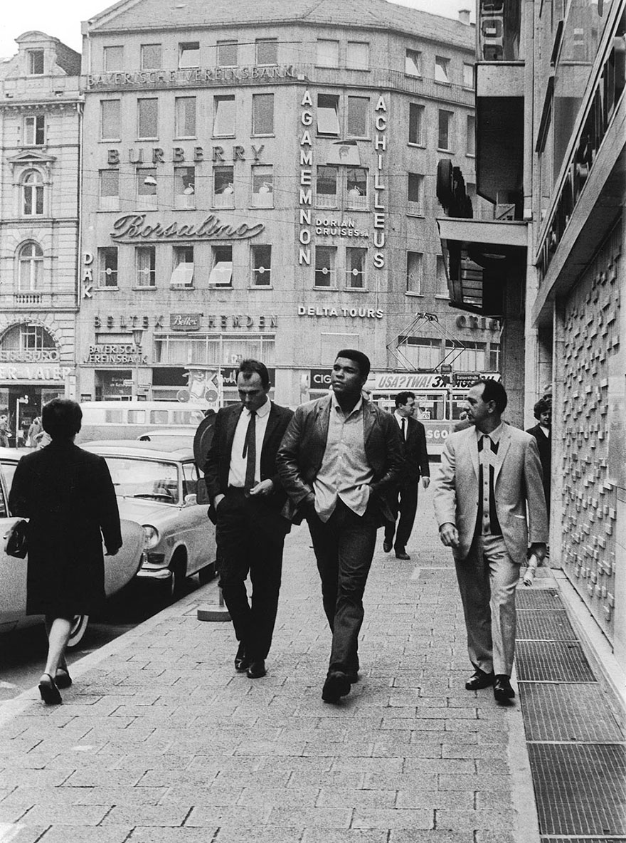 Muhammad Ali Walks Down A Main Street In Frankfurt, Germany With Boxing Promoter And Cornerman Angelo Dundee, While A Local Newsman Attempts To Get An Interview With The Prized Fighter, 1966