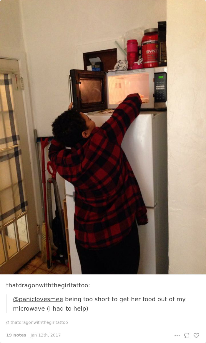 Person in a red plaid shirt reaching for food in a microwave on top of a fridge, highlighting short people problems.