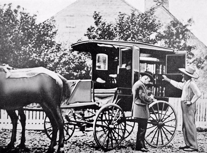 Early black and white photo of a horse-drawn bookmobile serving as mobile library with people selecting books outside.