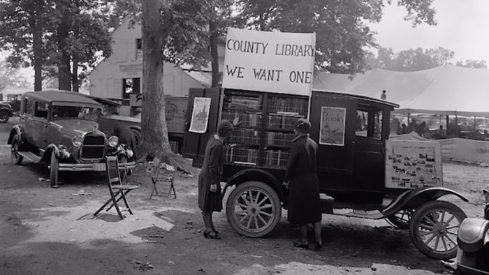 Two women browsing books from a vintage bookmobile parked outdoors, promoting county library services.
