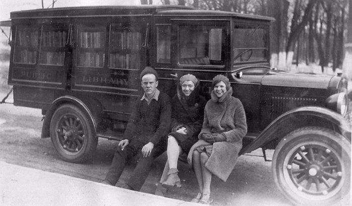 Three people sitting beside a vintage bookmobile, an early example of libraries-on-wheels providing mobile book access.